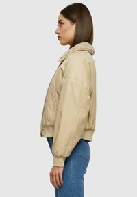Giacca bomber beige con un colletto testurizzato e morbido. Presenta un design aderente, polsini elasticizzati e tasche laterali, realizzata in tessuto liscio.