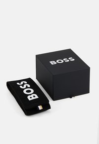 Boîte cadeau noire avec "BOSS" en lettres blanches, à côté d'une pochette en daim noir avec la même marque. Design compact et rectangulaire.