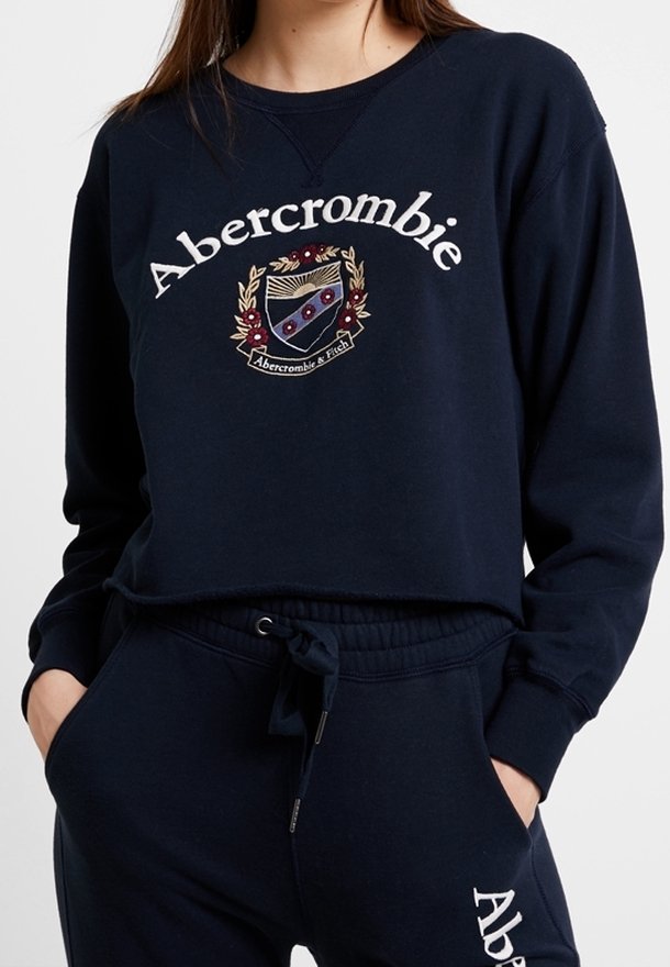Sudadera de color navy con el texto bordado "Abercrombie" y un diseño de emblema. Cuenta con cuello redondo y corte relajado, combinada con pantalones a juego.