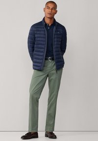 Veste matelassée bleu marine avec fermeture éclair, portée sur un polo bleu marine. Pantalon vert olive et chaussures marron foncé polis complètent la tenue.
