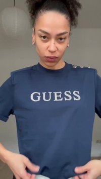 Göndör hajú nő, aki hátrafogott hajjal tengerészkék GUESS pólót visel, a ruhája alját igazítja meg, beltéren, egyszínű háttér előtt áll.