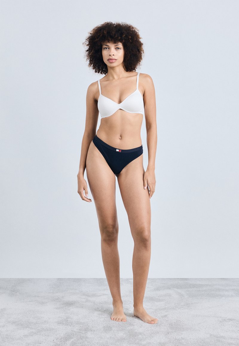 Bjela podstavljena grudnjak i crni bikini donji dio s logom, glatka tekstura i prilagođeni dizajn. Model stoji na sivoj pozadini.