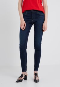 Vaqueros ajustados de denim azul oscuro con una textura suave, que cuentan con un diseño de cinco bolsillos y pasadores para cinturón, combinados con zapatos negros de tacón.