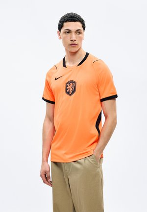 Νέος άνδρας που φοράει πορτοκαλί φανέλα ποδοσφαίρου με μαύρο έμβλημα λέοντα, στέκεται με το ένα χέρι στην τσέπη, κοιτάζοντας προς τα πλάγια.