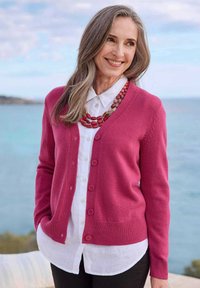 Rosa Strickjacke mit Knopfverschluss und geripptem Saum, über einem weißen Hemd mit Kragen drapiert. Auffällige Halskette mit roten Perlen und strukturiertem Material.