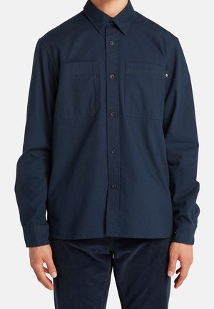 Mann trägt ein dunkelblaues Hemd mit langen Ärmeln und zwei Brusttaschen, steht vor einem schlichten hellen Hintergrund.