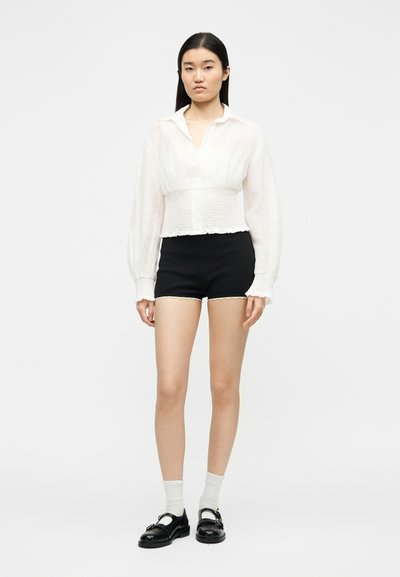 Femme debout portant un chemisier blanc à manches longues, un short taille haute noir, des chaussettes blanches et des chaussures Mary Jane noires, sur un fond uni.