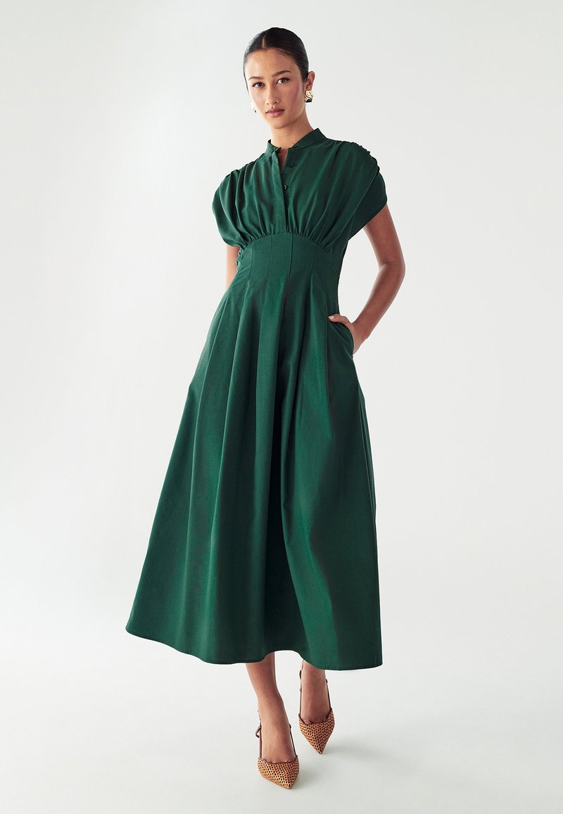 Žalia, iki kelių suknele su trumpomis pūstomis rankovėmis, prigludusia liemene ir raukšlėta sijonine dalimi. Kišenės abiejose pusėse. Dėvima su smailiais smėlio spalvos aukštakulniais.