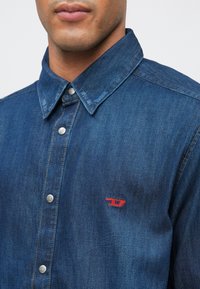 Άνδρας που φοράει ένα μπλε τζιν πουκάμισο με κουμπιά και σκληρό γιακά, με ένα μικρό κόκκινο ραμμένο λογότυπο στο στήθος.