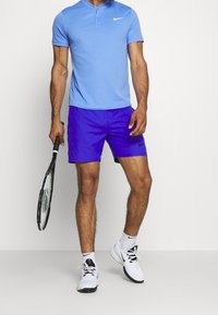 Camisa de collar de manga corta azul y pantalones cortos morados, ambos con el logo de Nike. Zapatillas de tenis blancas con suelas negras. Sosteniendo una raqueta de tenis.