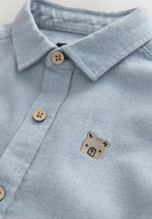Light blue collared shirt with textured fabric, featuring wooden buttons and a small embroidered bear graphic on the chest.