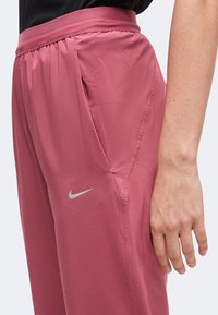 Άτομο που φοράει φαρδιά ροζ παντελόνια Nike και μαύρη μπλούζα, με το δεξί χέρι στο πλάι, δείχνοντας το λογότυπο της Nike κοντά στην τσέπη.