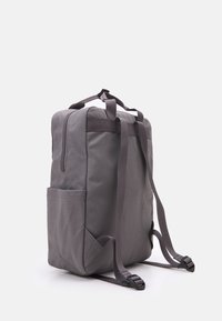 Sac à dos en tissu gris, de forme rectangulaire, avec des bretelles réglables, une poche avant et un compartiment zippé. Texture lisse et design minimal.