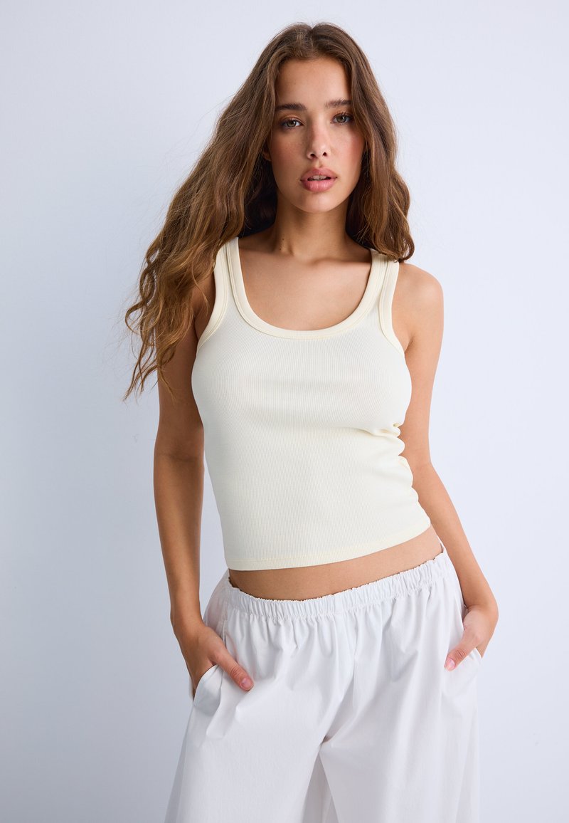 Jeune femme aux longs cheveux ondulés portant un haut crème sans manches et un pantalon blanc ample, debout les mains dans les poches devant un fond uni.