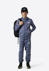 Plavi atletski trenirka s bijelim logotipom, jakna na patentni zatvarač i hlače. Crna kapa i ruksak upotpunjuju odjeću. Tenisice s bijelim potplatom.