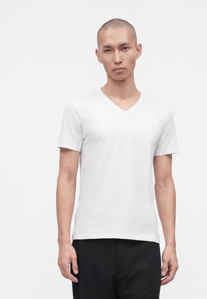 Homme à la tête rasée portant un t-shirt blanc uni à col en V et manches courtes, ainsi qu’un pantalon noir, sur fond clair uni.
