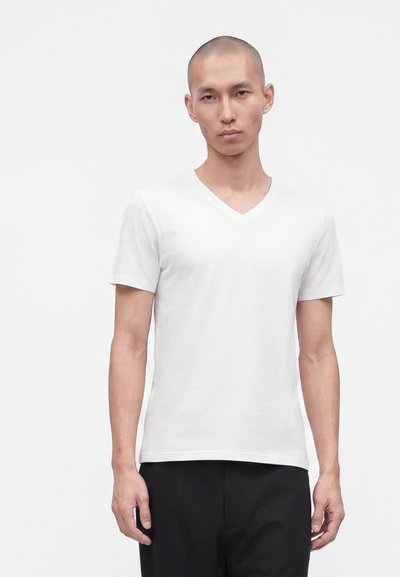 Homme à la tête rasée portant un t-shirt blanc uni à col en V et manches courtes, ainsi qu’un pantalon noir, sur fond clair uni.