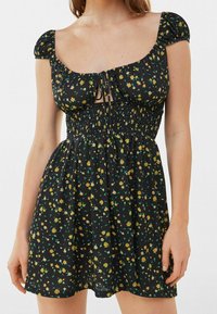 Schwarzes Blumenkleid mit kurzen Puffärmeln, gerafftem Oberteil und quadratischem Ausschnitt. Verfügt über ein Bindeband vorne und einen fließenden Rock.