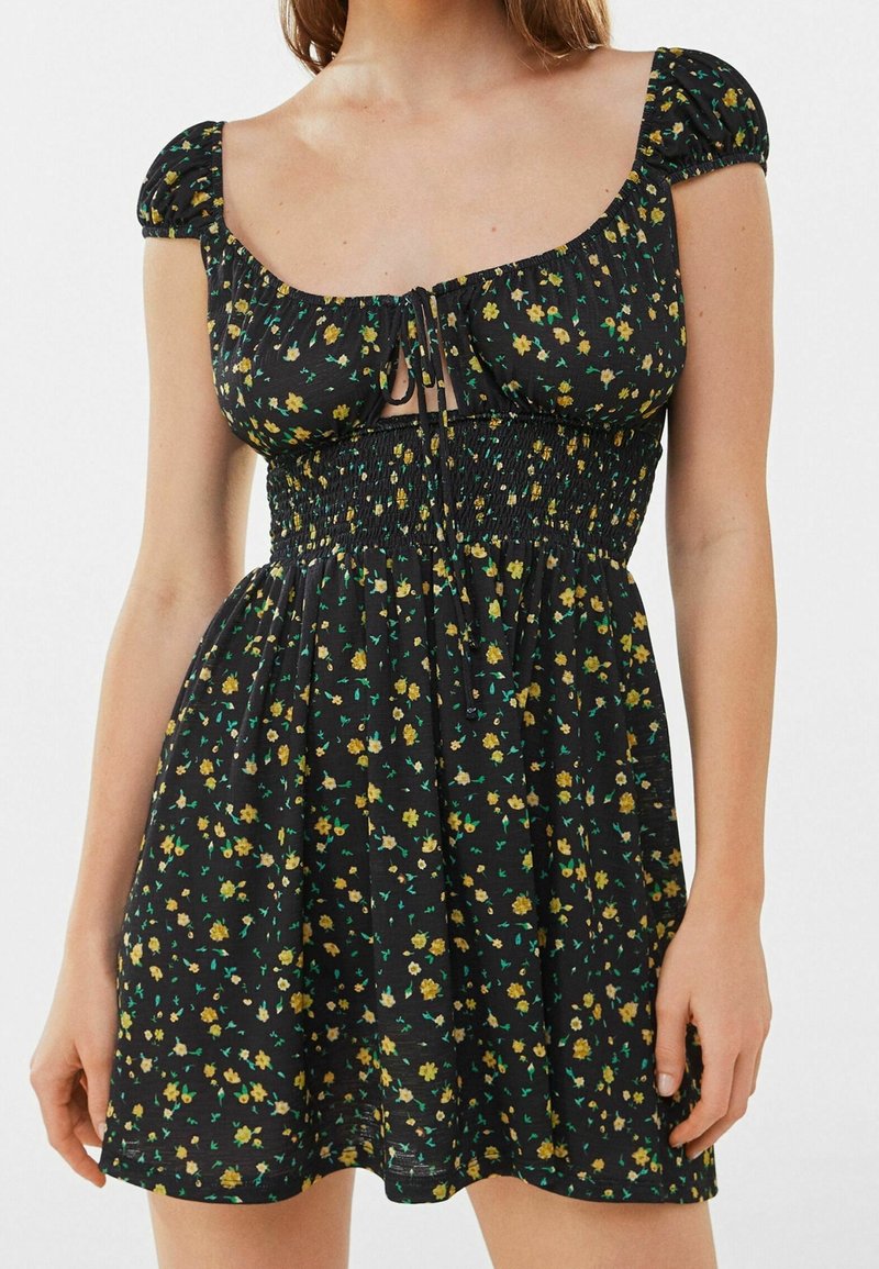Schwarzes Blumenkleid mit kurzen Puffärmeln, gerafftem Oberteil und quadratischem Ausschnitt. Verfügt über ein Bindeband vorne und einen fließenden Rock.
