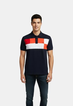 Hombre vistiendo una camiseta tipo polo azul marino con bloques de color rojo, naranja y blanco en el pecho y jeans azul oscuro sobre un fondo liso.