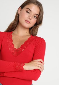 Maglietta rossa a maniche lunghe con scollatura a V e bordi in pizzo. Presenta un design aderente e una texture a coste, accentuando uno stile casual.