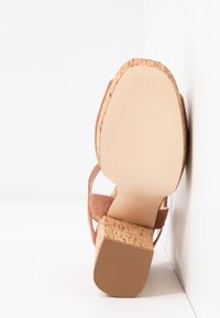 Sandačky s korkovým blokovým podpatkem a semišovými popruhy umístěné podrážkou nahoru proti bílým stěnám, zdůrazňující hladký a texturovaný podešev.