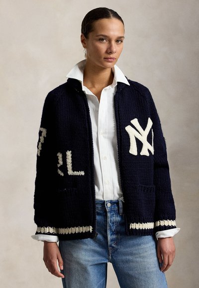 Femme portant une veste en maille bleu marine avec des lettres blanches "NY", une chemise blanche à col et un jean bleu, debout devant un fond neutre uni.