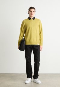 Gul genser i strikk, svart skjorte med krage under, svarte bukser, hvite sneakers og en svart skulderveske; uformell antrekk med avslappet passform.