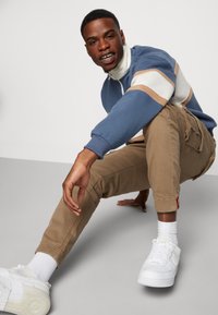 Blaues und cremefarbenes Zip-up Sweatshirt mit Streifen, kombiniert mit hellbraunen Cargo-Hosen und weißen Sneakers. Glattes Material, lockere Passform.
