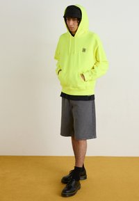 Neon yellow hoodie with drawstrings and front pockets, paired with gray shorts. Black shoes with thick soles, black socks, and a black cap.
