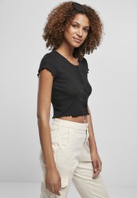 Top noir côtelé à manches courtes à volants et détails de boutons, associé à un pantalon crème avec poches latérales.