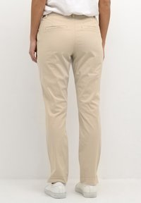Beige cotton blend trousers with a straight leg, featuring back pockets and stitched detailing, paired with white low-top shoes.