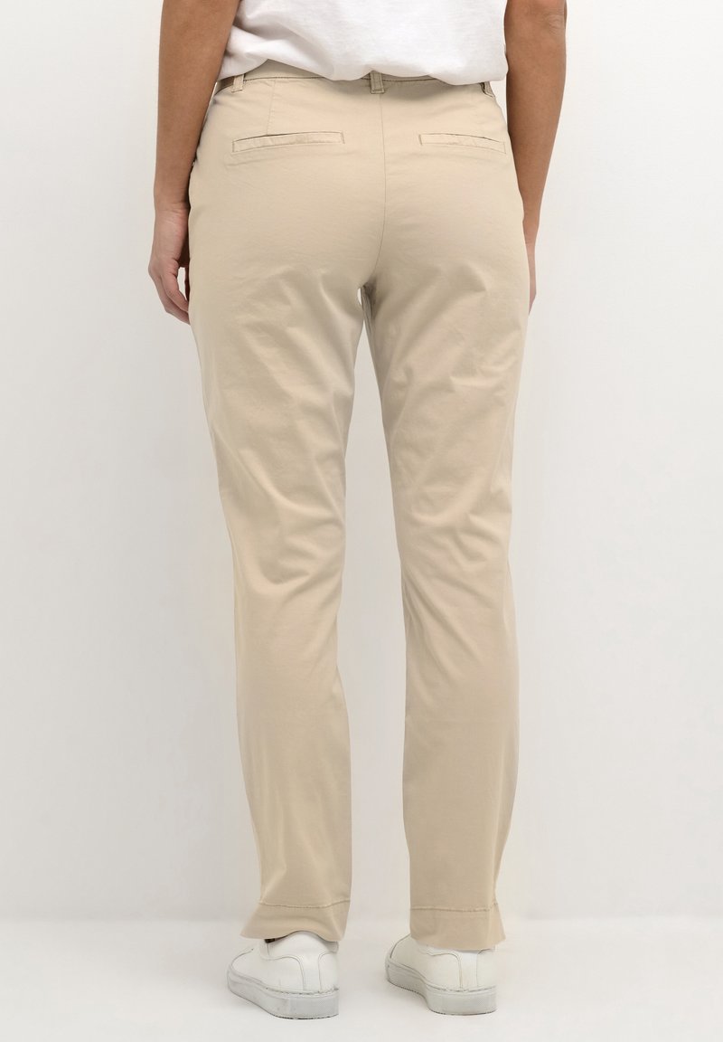 Beige cotton blend trousers with a straight leg, featuring back pockets and stitched detailing, paired with white low-top shoes.
