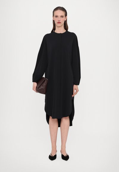 Vestido preto de mangas longas com um ajuste solto, decote redondo, fendas laterais e comprimento até ao joelho, combinado com uma bolsa castanha escura.