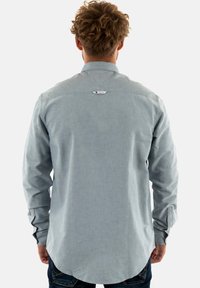 Camisa de manga larga azul claro con cuello abotonado, detalle en la espalda y dobladillo curvado. Hecha de tela texturizada, incluye una pequeña etiqueta con logo.