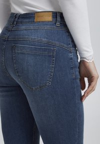 Šviesiai mėlyni džinsai, turintys aukštą liemenį, nugarinius kišenes su siuvinėjimo akcentais ir odinį prekės ženklo etiketę ant juosmens.