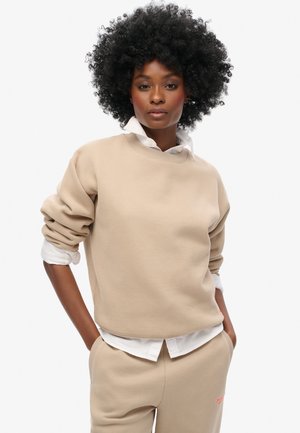 Felpa beige con scollo rotondo e maniche arrotolate, indossata sopra una camicia bianca. Presenta una texture morbida e un sottile dettaglio di branding.