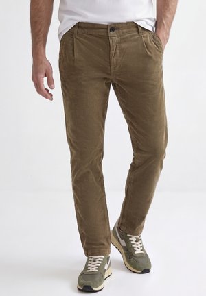 Homme portant un pantalon en velours côtelé vert olive et des baskets gris-vert avec des lacets blancs, debout avec une main dans la poche.