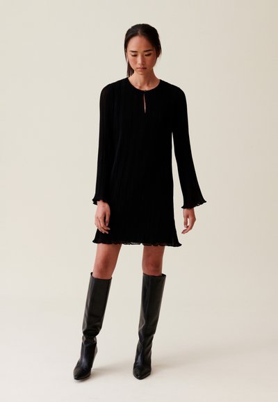 Robe noire plissée à manches longues, encolure avec ouverture, ourlet festonné et coupe décontractée, assortie à des bottes en cuir noir montantes.