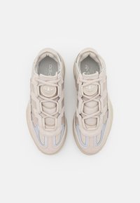 Zapatillas de deporte color beige claro con parte superior texturizada, detalles de ante y elementos reflectantes. Presenta un logo redondo y un sistema de cordones elásticos.