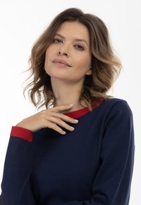 Cămașă lungă de culoare albastru marin, cu guler și mansete roșii. Materialul pare moale și texturat. Modelul își sprijină mâna pe gât.