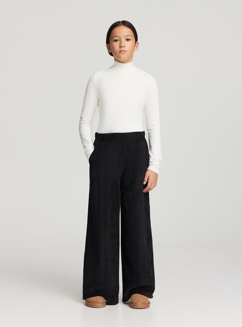 Top bianco a maniche lunghe con collo alto abbinato a pantaloni neri larghi in velluto a coste e scarpe slip-on color cognac. Design e texture semplici.
