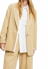 Personne portant un blazer en velours côtelé beige et un pantalon large assorti par-dessus une chemise blanche à boutons, posant devant un fond blanc.