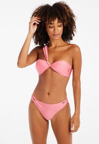 Růžový bikini set obsahující top na jedno rameno s uzlem a odpovídající bikini kalhotky s uzlem. Hladká textura s minimálními detaily kovových prvků.