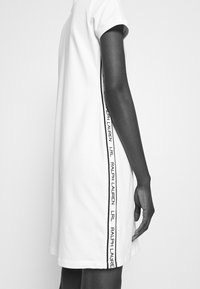 Vit bomullsklänning med korta ärmar, prydd med svarta sidosträck med texten "LRL RALPH LAUREN." Enkel design och avslappnad passform.