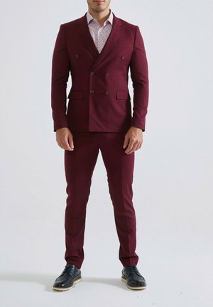 Homme portant un costume bordeaux ajusté à double boutonnage avec un pantalon assorti, une chemise blanche à motifs et des chaussures habillées noires, debout devant un fond uni.