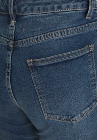 Os jeans em denim azul escuro apresentam um bolso traseiro com detalhes de costura e uma textura suave. O tecido parece durável e ajustado ao corpo.