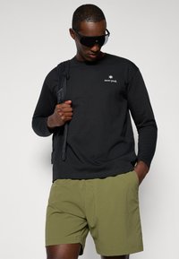 Black long-sleeve shirt with white logo, paired with light olive green shorts. Model wears black sunglasses and carries a black backpack.