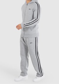 Grauer Hoodie und passende Jogginghose mit schwarzen Seitenstreifen. Kapuzen-Design, Reißverschluss und Seitentaschen. Weiße Schuhe.