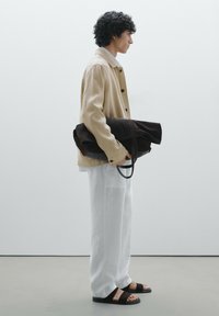 Veste beige, chemise gris clair, pantalon blanc, sandales noires. Tenant un sac en daim marron foncé avec un dessus enroulé et des poignées noires.
